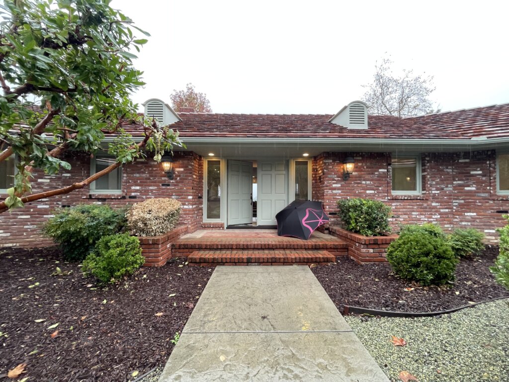 Rainy day Fallbrook open house front exterior of 145 Yucca Road with brick entry, porch lights, and umbrella by the door. 